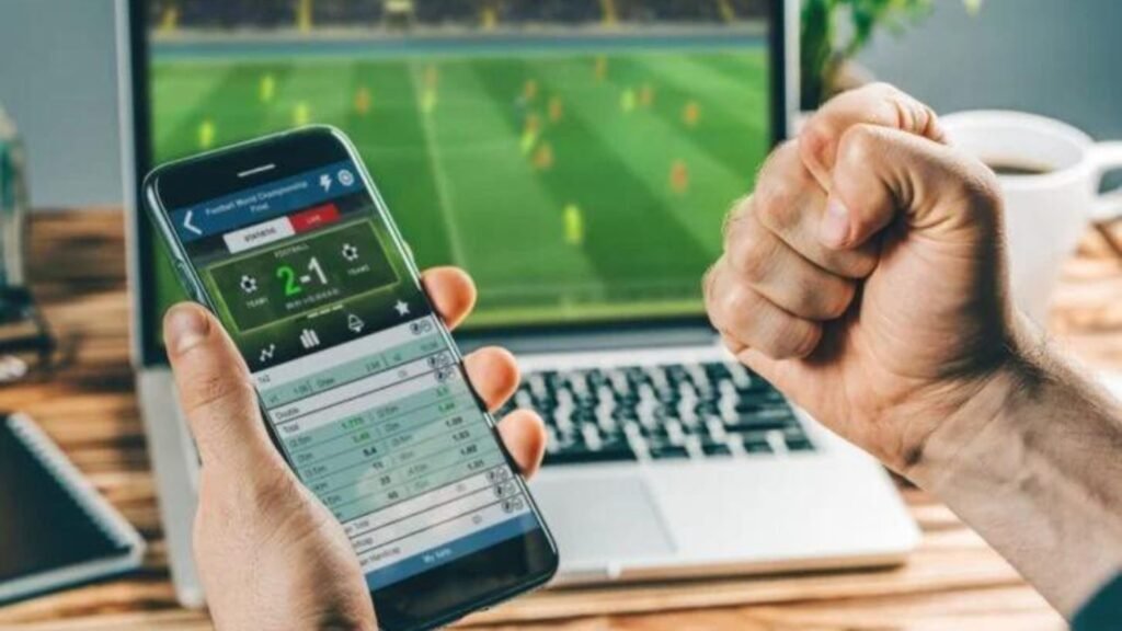 a man holding a phone with a laptop in front showing Online Betting vs. Traditional Betting